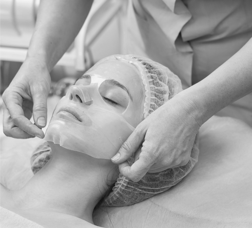 Image monochrome montrant les mains d'une esthéticienne appliquant un masque sur le visage d'une cliente.